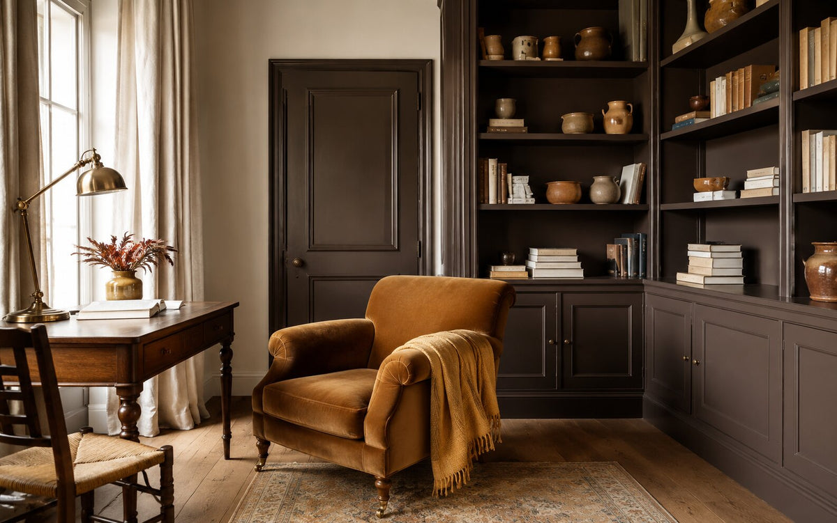 Studeerkamer met een diep chocoladebruine boekenkast en kozijn, een caramelkleurige velours fauteuil met honingkleurige plaid, en warme gebroken witte muren met linnen gordijnen