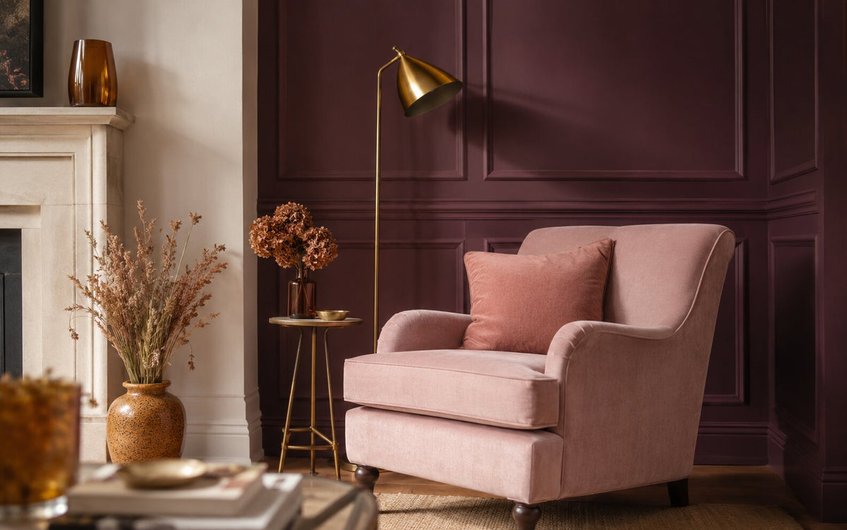 Sfeervolle zithoek met een diepe pruim/aubergine gelambriseerde wand, een poederroze velours fauteuil met dusky-rose kussen, een messing vloerlamp en bijzettafel, een gebrande okerkleurige vaas en een crème schouw