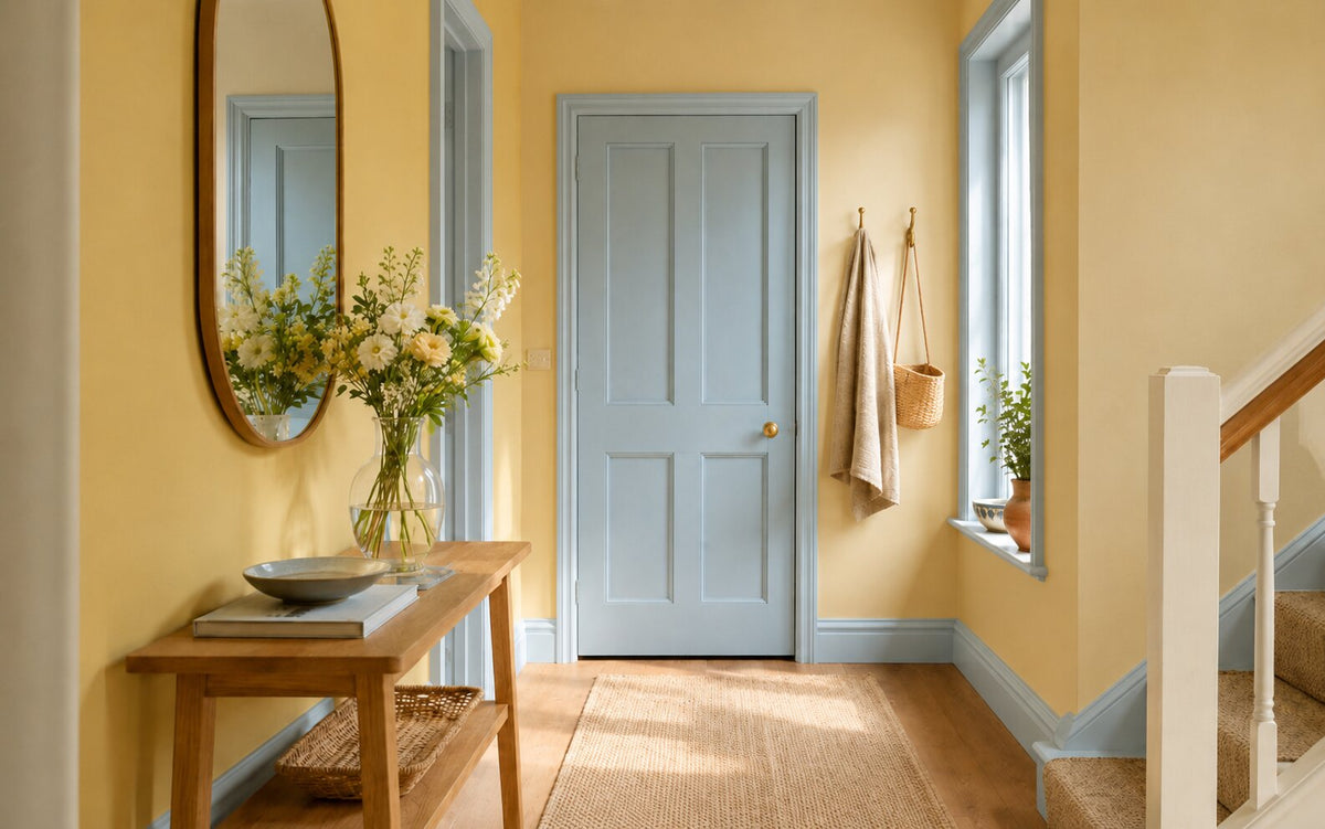 Lichte hal met alle muren in een zacht geel, de paneeldeur, plint en kozijnen in een licht poederblauw, een eiken sidetable met spiegel en verse bloemen, en een jute loper