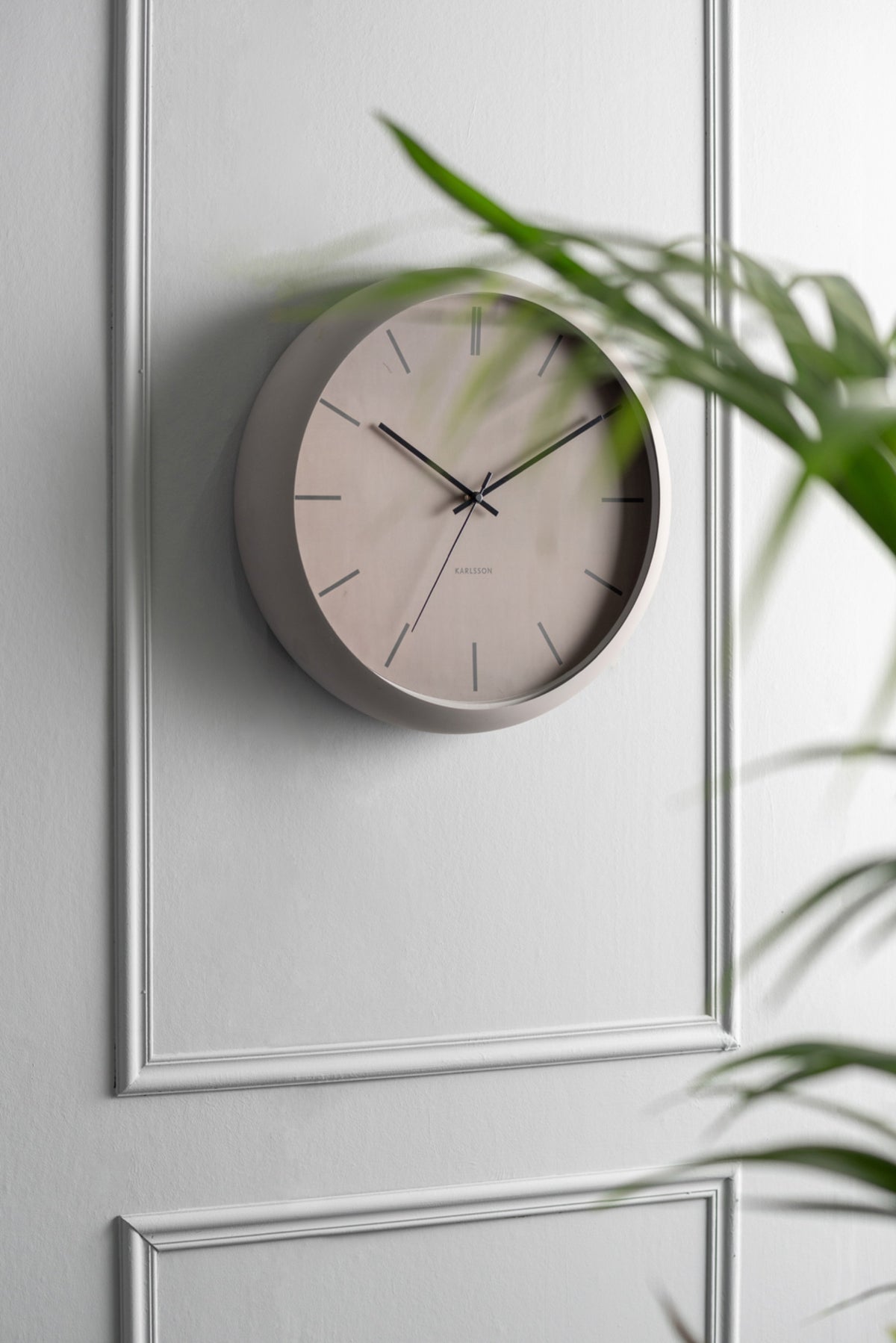 Karlsson wandklok Nirvana Globe, warm grijs, sfeerfoto: schuin aan witte lambriseringwand met palmblad