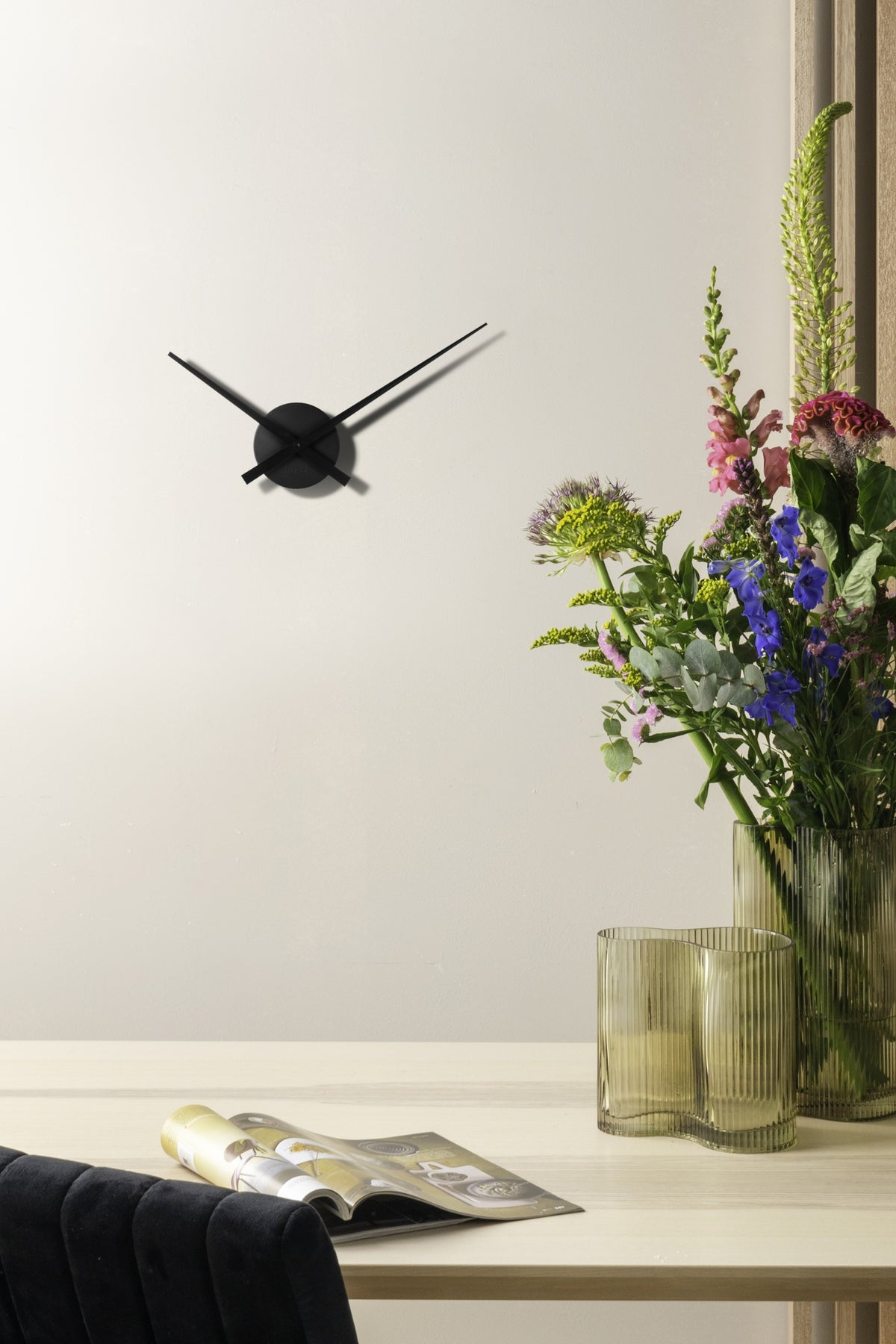 Karlsson wandklok Little Big Time Mini, zwart aluminium, sfeerfoto: aan lichte wand naast bloemen in geribbelde vazen