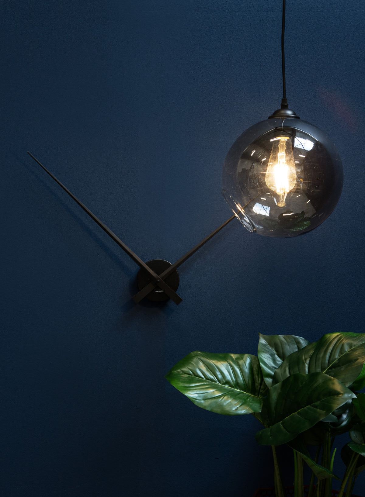 Karlsson wandklok Little Big Time, zwart aluminium, sfeerfoto: aan donkerblauwe wand met hanglamp en plant