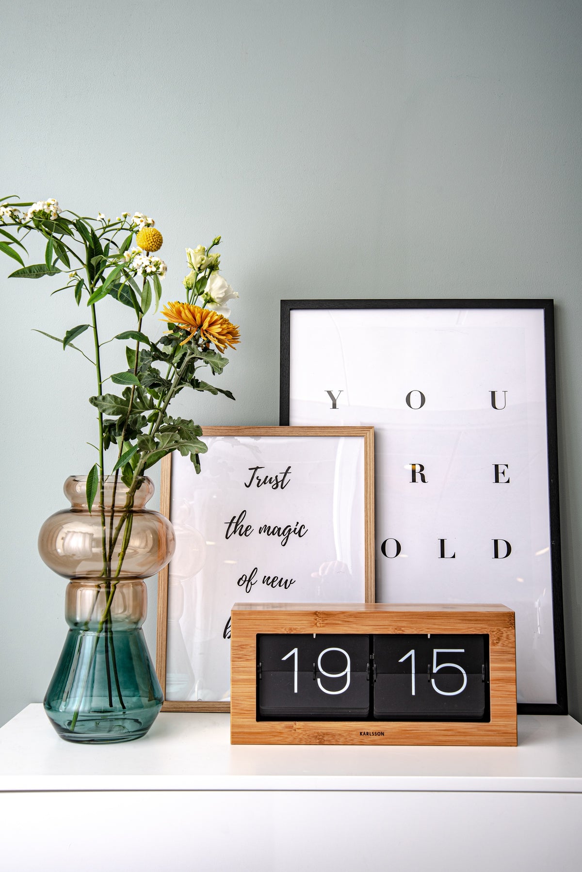 Karlsson flipklok Boxed XL, bamboe, sfeerfoto: op wit dressoir voor posters en vaas met bloemen, toont 19:15