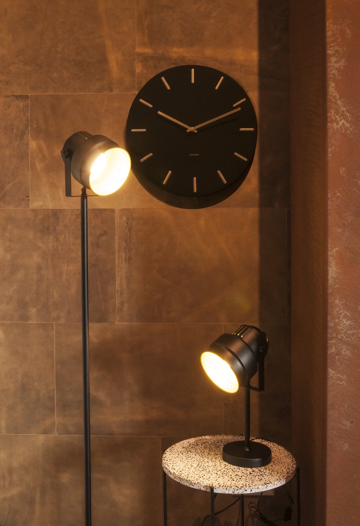 Karlsson wandklok Charm, zwart, sfeerfoto: aan bruine wand met twee zwarte spotlampen en terrazzo bijzettafel