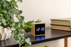Karlsson wekker Mini Cube, bamboe, sfeerfoto: op zwart bureau naast klimop en boeken, display toont 12:10