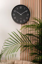 Karlsson wandklok Lofty, ijzer mat zwart, sfeerfoto: aan lichte wand naast houten lattenwand met palm