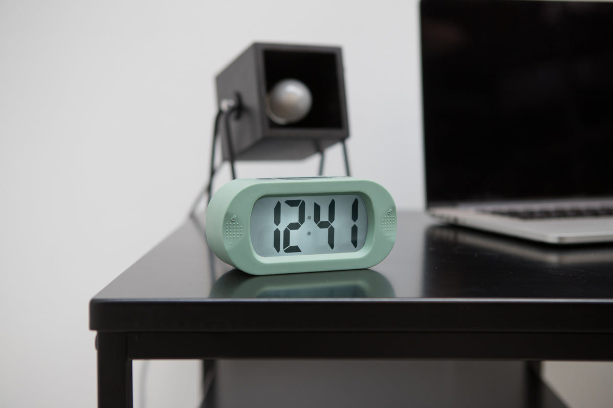Karlsson wekker Gummy Rubberized, groen, sfeerfoto: op zwart bureau naast laptop en lamp, display toont 12:41