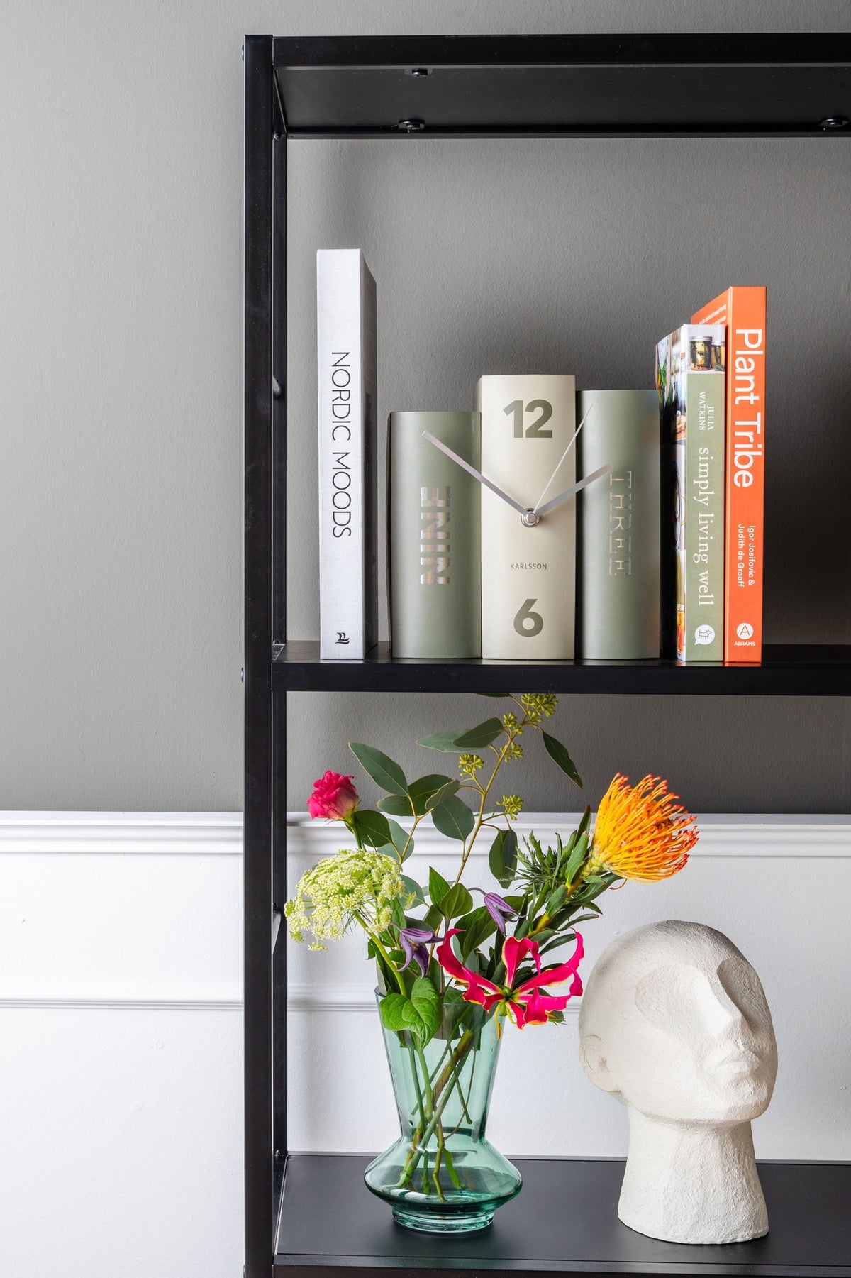 Karlsson tafelklok Book, groen paper, sfeerfoto: tussen boeken op zwart stellingrek, bloemen en beeld eronder