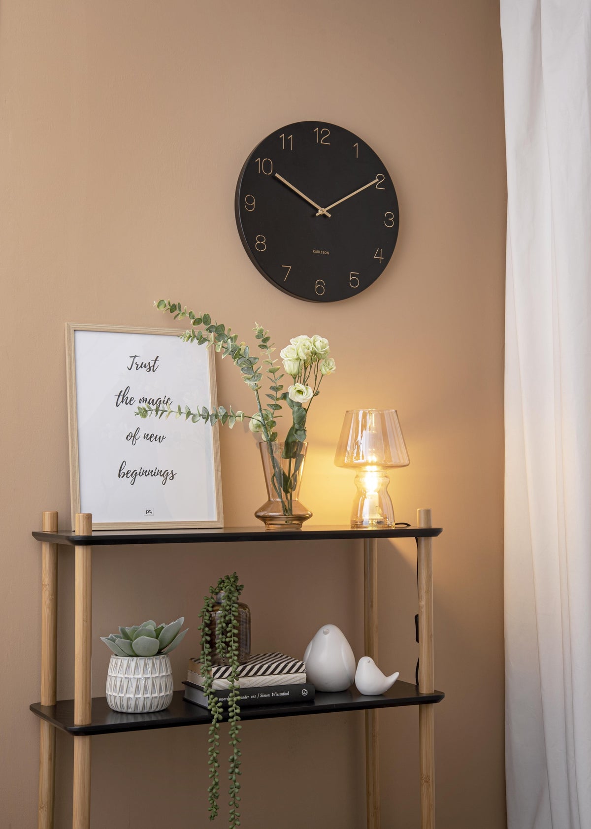Karlsson wandklok Charm, gegraveerde nummers, zwart, sfeerfoto: aan beige wand boven zwart wandrek met lamp