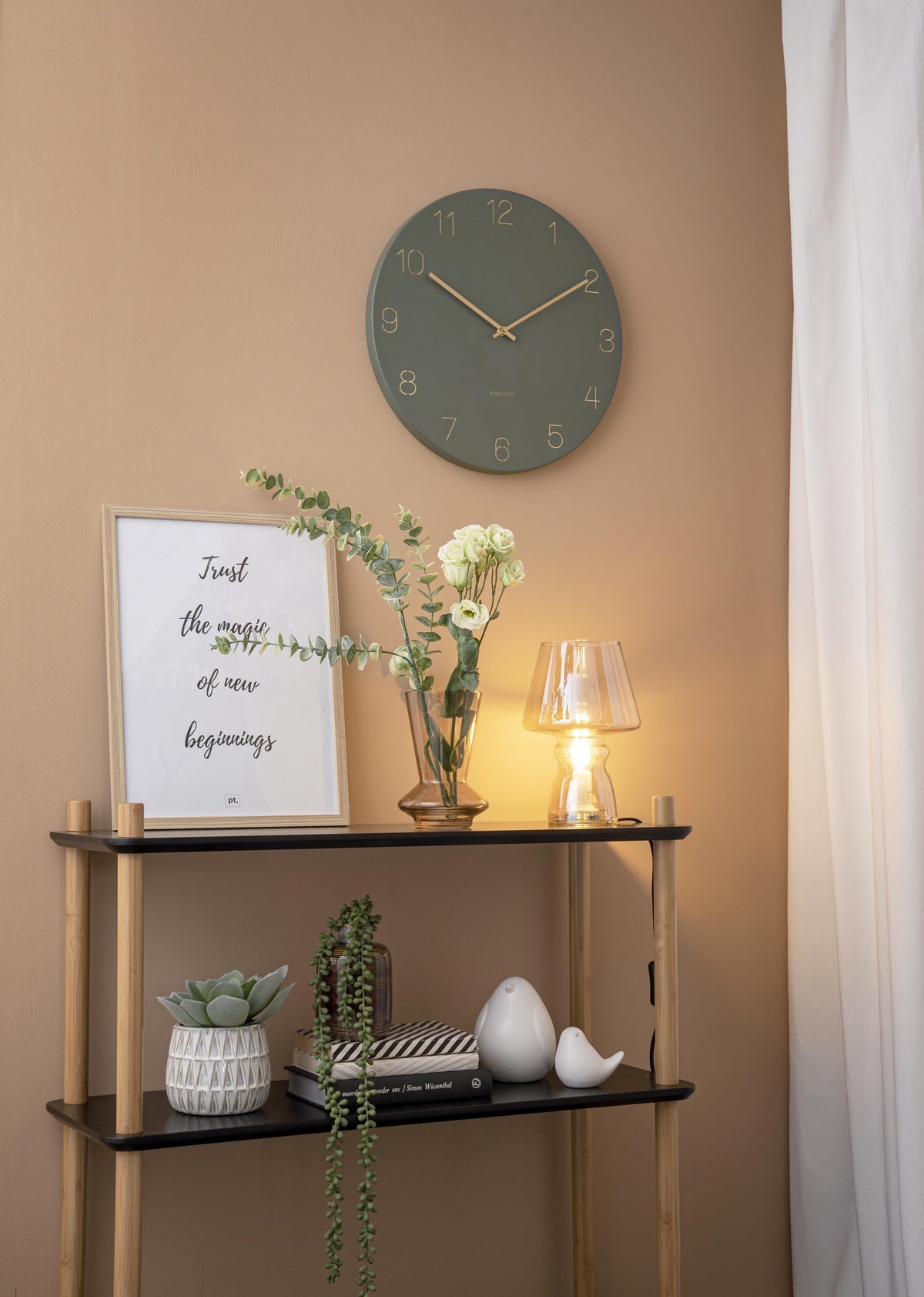 Karlsson wandklok Charm, gegraveerde nummers, jungle groen, sfeerfoto: aan beige wand boven zwart wandrek met lamp
