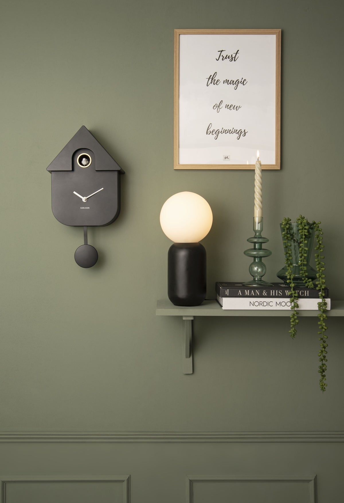 Karlsson koekoeksklok Modern Cuckoo, zwart abs, sfeerfoto: aan groene muur naast wandplank met lamp en kaars