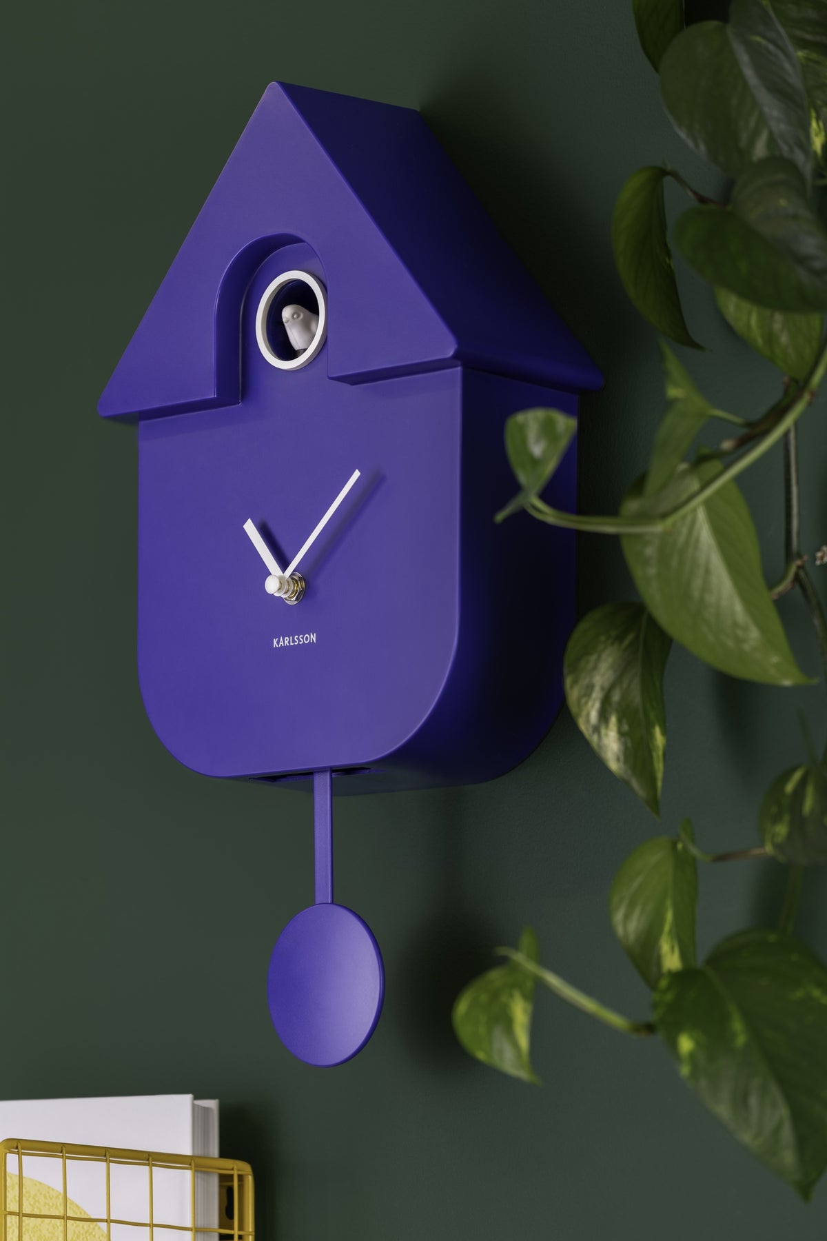 Karlsson koekoeksklok Modern Cuckoo, kobaltblauw, sfeerfoto: aan donkergroene wand naast hangplant, geel draadrek onderin