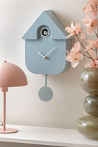 Karlsson koekoeksklok Modern Cuckoo, lichtblauw abs, sfeerfoto: schuin aan wand naast roze lamp en vaas met bloesem