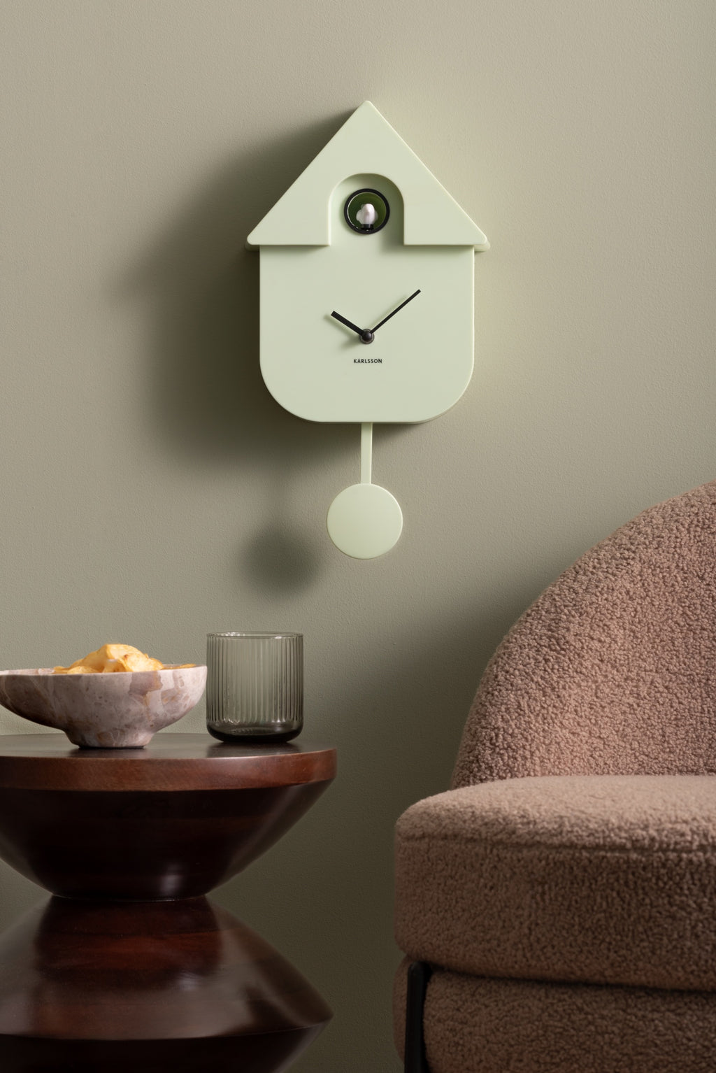 Karlsson koekoeksklok Modern Cuckoo, zachtgroen, sfeerfoto: aan groengrijze muur naast bruine fauteuil en bijzettafel