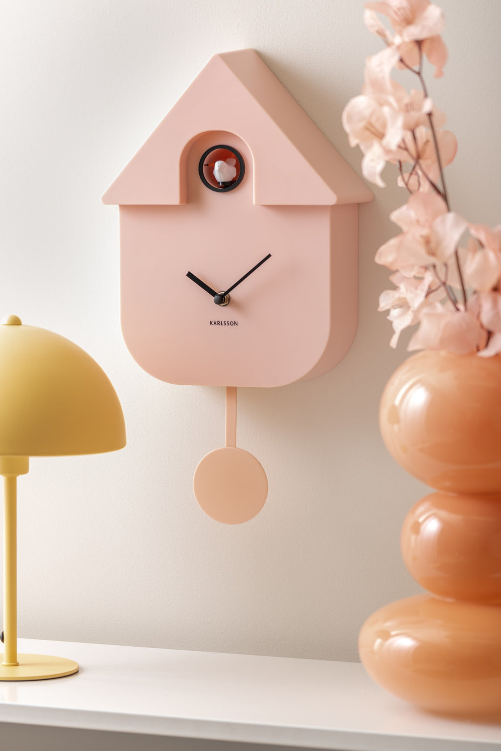 Karlsson koekoeksklok Modern Cuckoo, zachtroze abs, sfeerfoto: schuin aan wand naast gele lamp en oranje vaas met bloesem