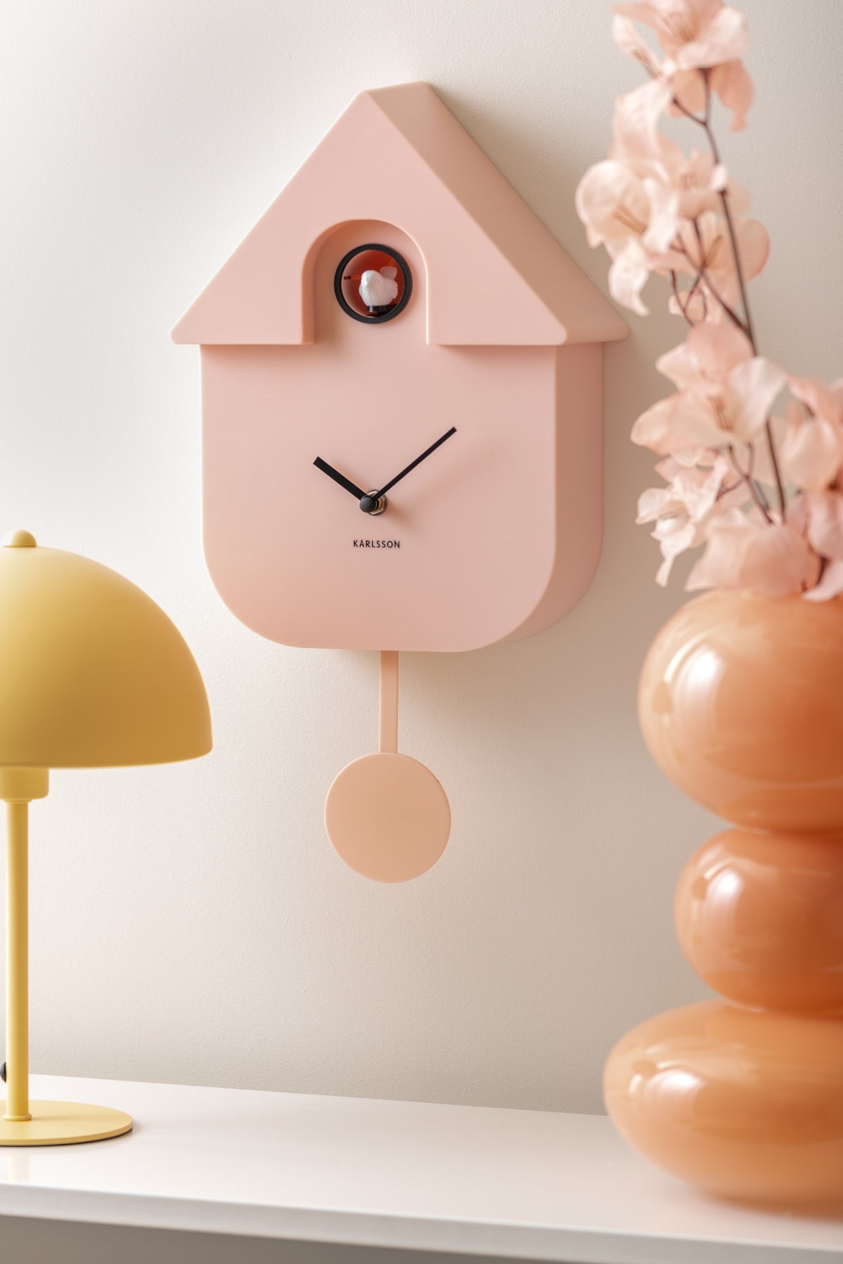 Karlsson koekoeksklok Modern Cuckoo, zachtroze abs, sfeerfoto: schuin aan wand naast gele lamp en oranje vaas met bloesem