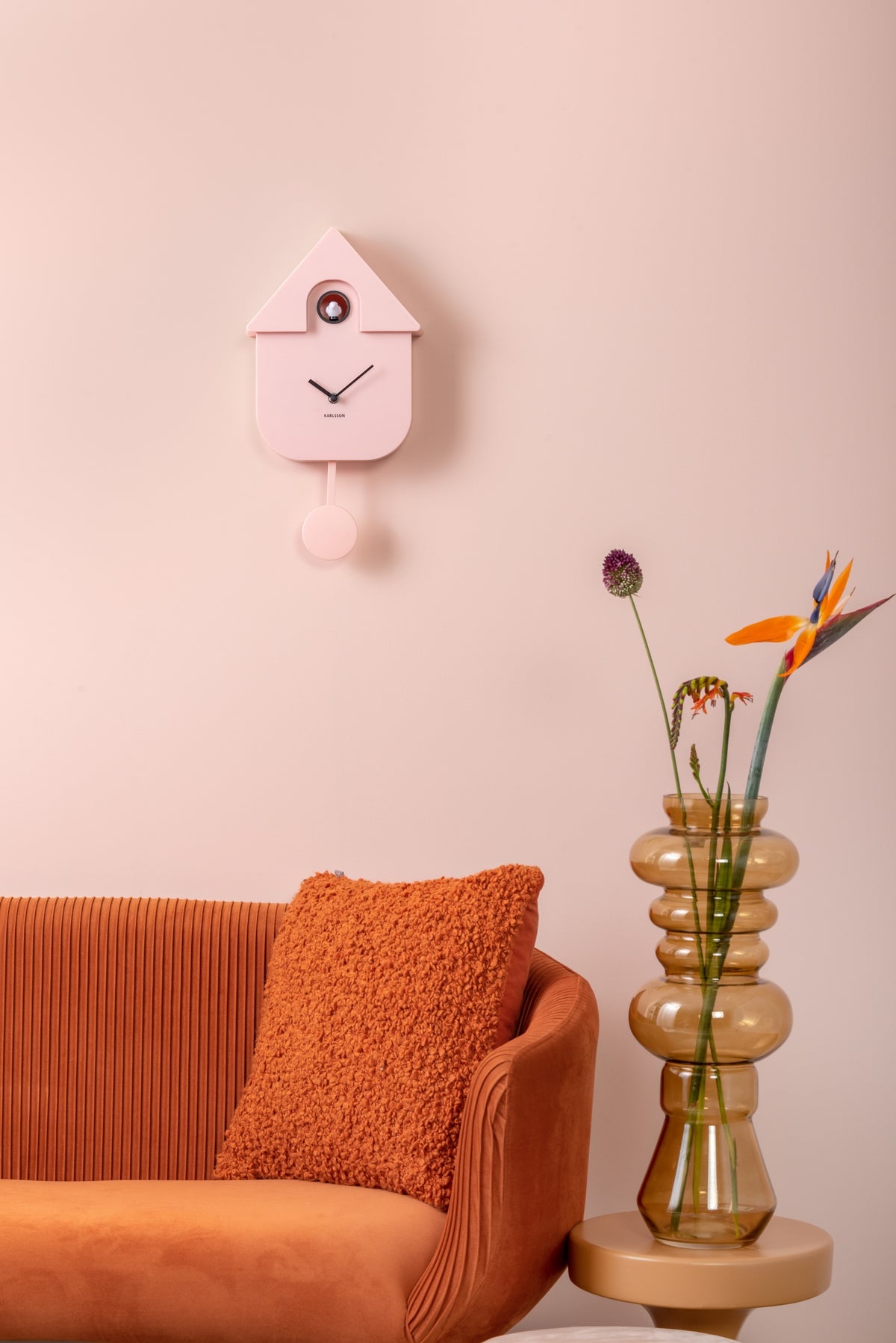 Karlsson koekoeksklok Modern Cuckoo, zachtroze abs, sfeerfoto: aan roze wand boven oranje bank met vaas en bloemen