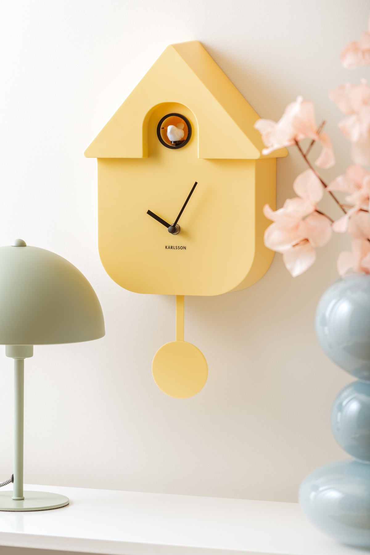 Karlsson koekoeksklok Modern Cuckoo, zachtgeel abs, sfeerfoto: schuin aan wand naast groene lamp en blauwe vaas met bloesem