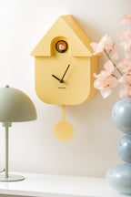 Karlsson koekoeksklok Modern Cuckoo, zachtgeel abs, sfeerfoto: schuin aan wand naast groene lamp en blauwe vaas met bloesem