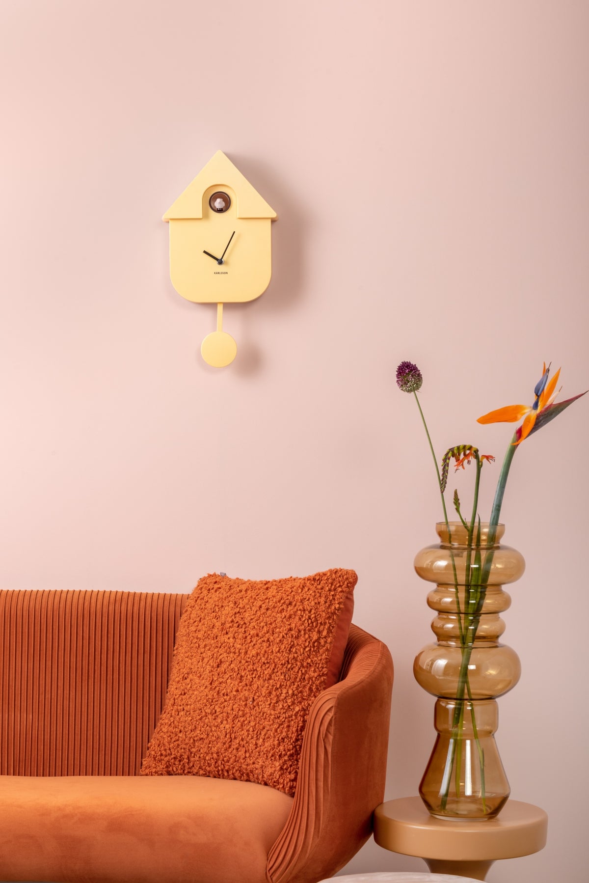 Karlsson koekoeksklok Modern Cuckoo, zachtgeel abs, sfeerfoto: aan roze wand boven oranje bank met vaas en bloemen