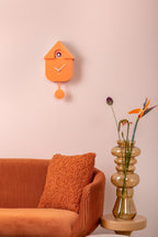 Karlsson koekoeksklok Modern Cuckoo, zachtoranje abs, sfeerfoto: aan roze wand boven oranje bank met vaas en bloemen