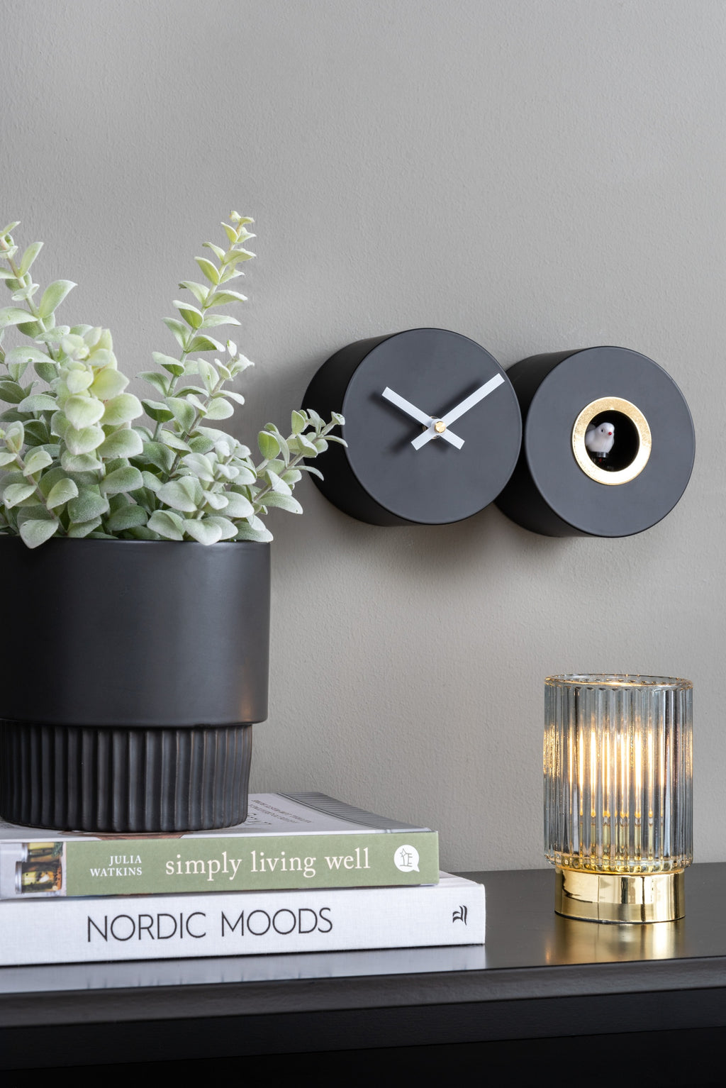 Karlsson koekoeksklok Duo Cuckoo, mat zwart, sfeerfoto: aan grijze wand naast plant, boeken en geribbelde lamp