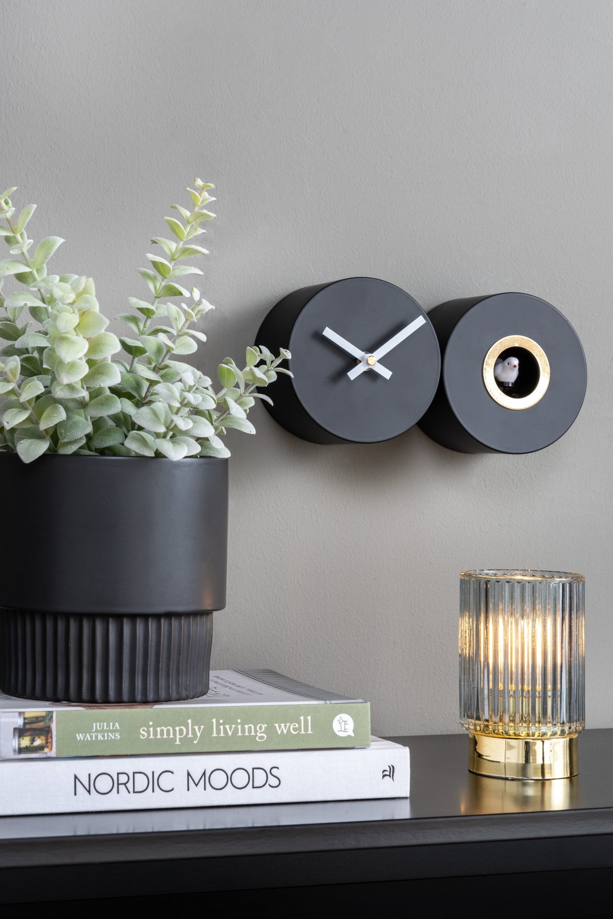 Karlsson koekoeksklok Duo Cuckoo, mat zwart, sfeerfoto: aan grijze wand naast plant, boeken en geribbelde lamp