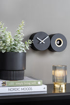 Karlsson koekoeksklok Duo Cuckoo, mat zwart, sfeerfoto: aan grijze wand naast plant, boeken en geribbelde lamp