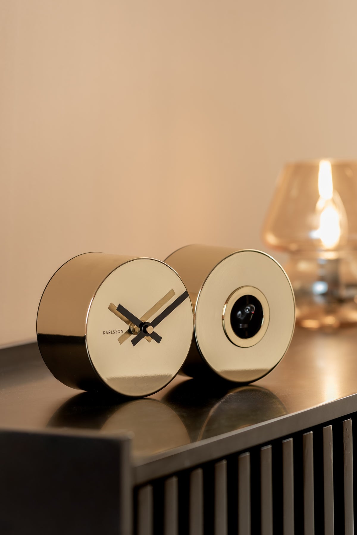 Karlsson koekoeksklok Duo Cuckoo, goud (verguld), sfeerfoto: op donker dressoir bij warme tafellamp