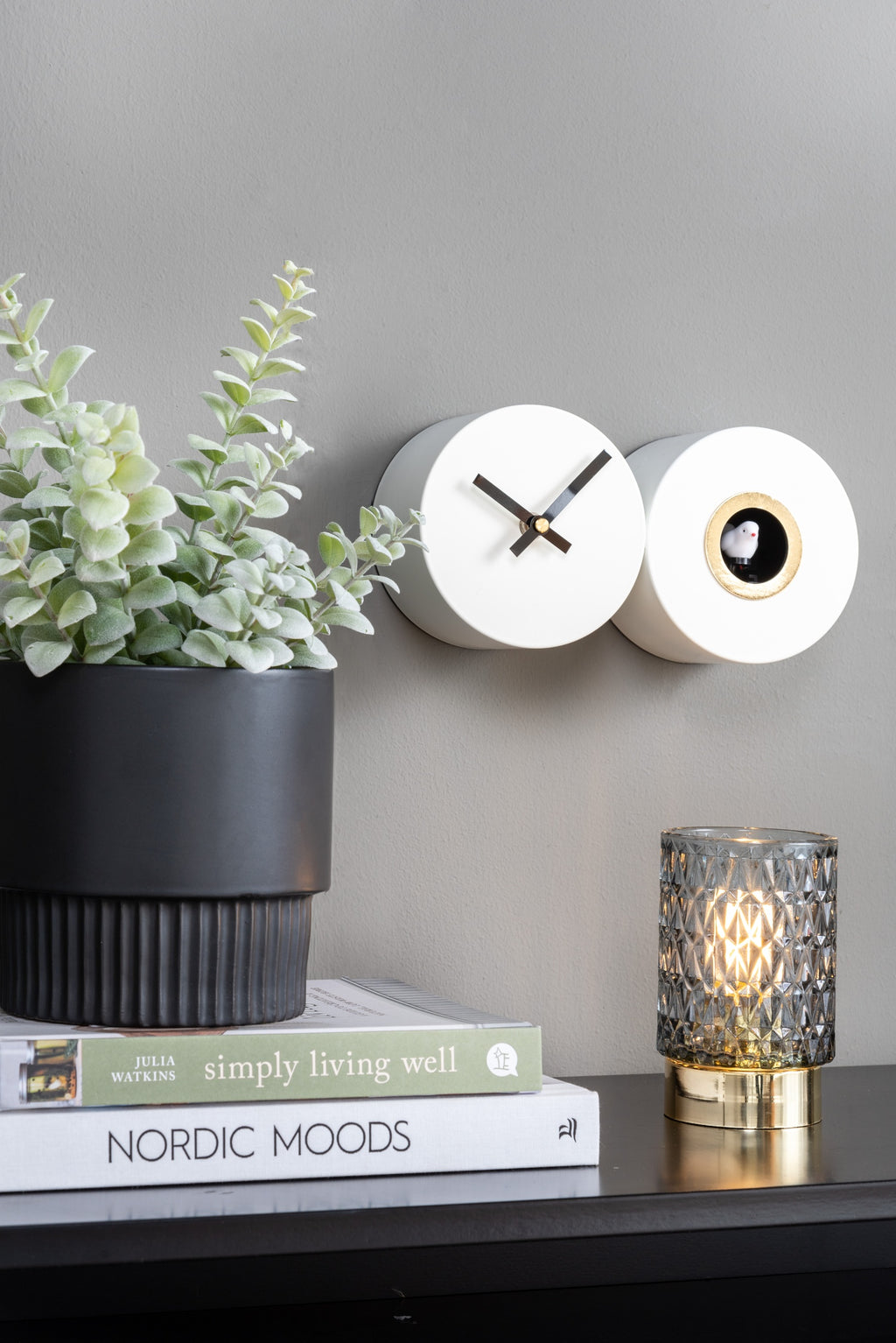 Karlsson koekoeksklok Duo Cuckoo, mat wit, sfeerfoto: aan grijze wand naast plant, boeken en geribbelde lamp