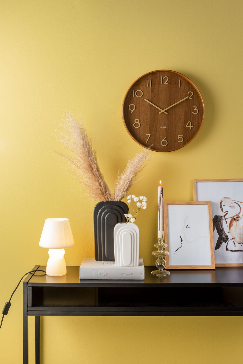 Karlsson wandklok Pure Medium, donker lindehout, sfeerfoto: aan gele wand boven zwarte sidetable met vazen