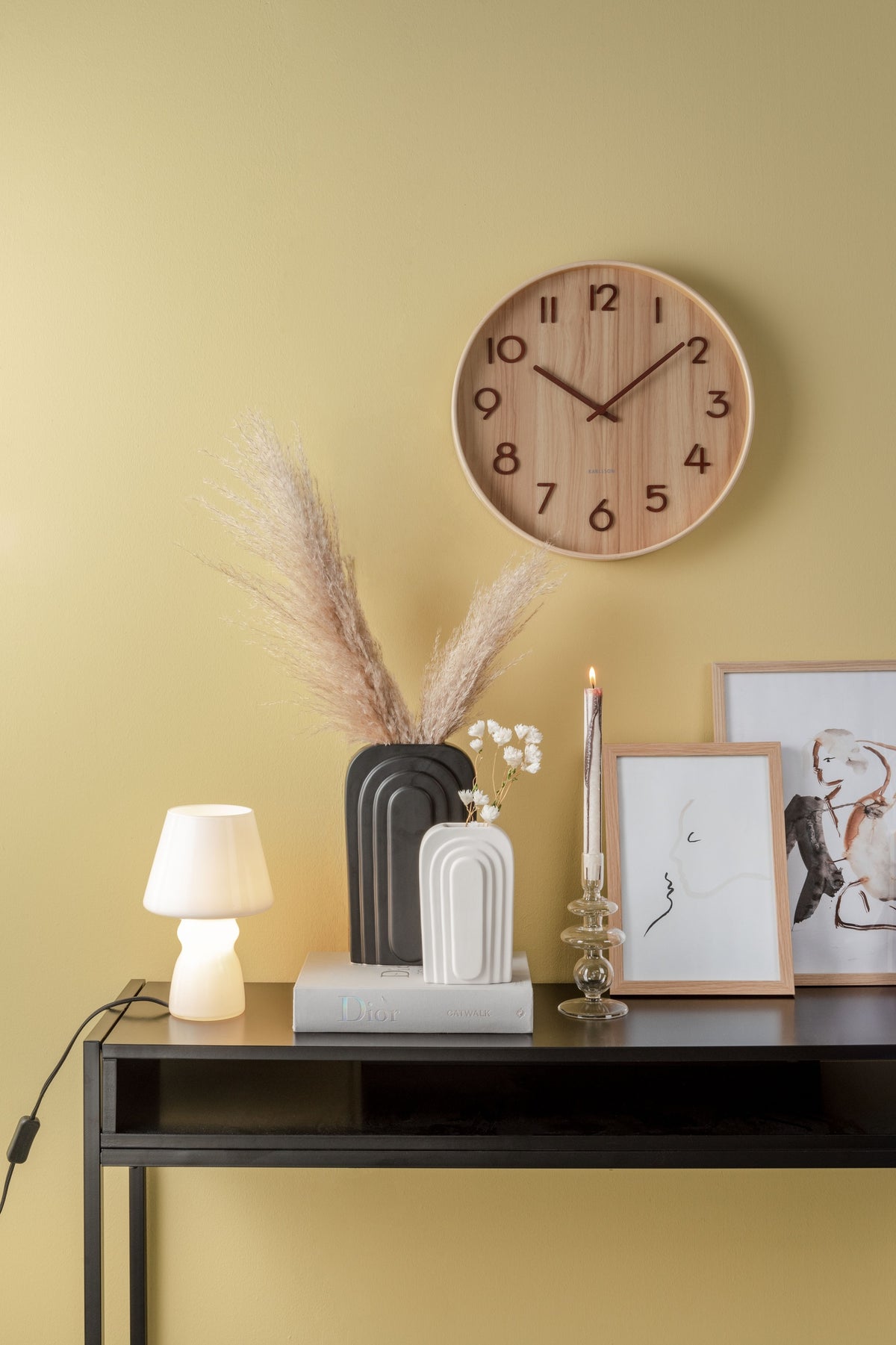 Karlsson wandklok Pure Medium, licht lindehout, sfeerfoto: aan gele wand boven zwarte sidetable met vazen, pluimgras en lamp