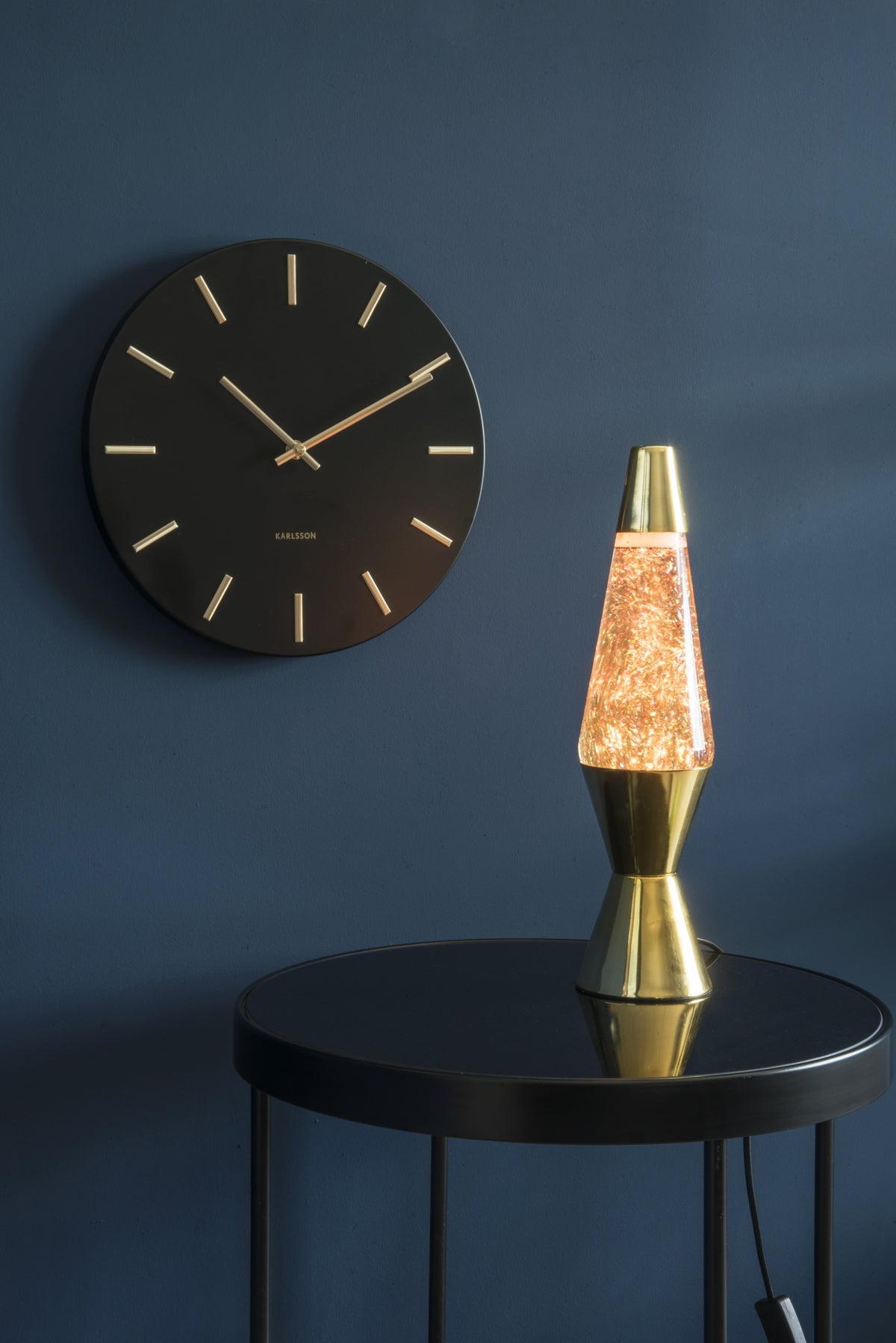 Karlsson wandklok Charm Small, zwart, sfeerfoto: aan donkerblauwe wand naast gouden lavalamp op zwart bijzettafeltje