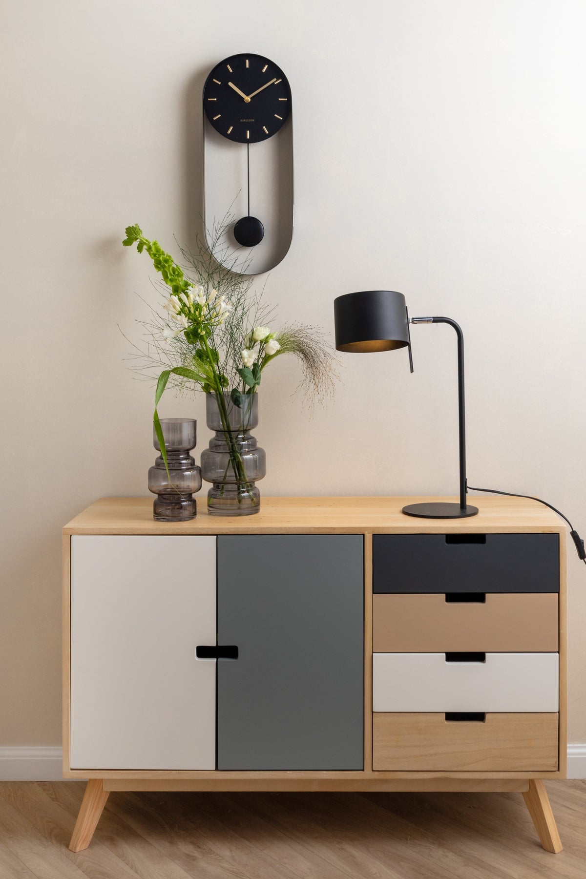 Karlsson wandklok Pendulum Charm, staal zwart, sfeerfoto: aan lichte wand boven dressoir met zwarte lamp en vazen