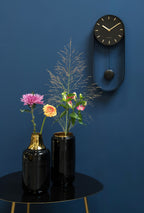Karlsson wandklok Pendulum Charm, staal zwart, sfeerfoto: aan donkerblauwe wand naast zwarte vazen met bloemen