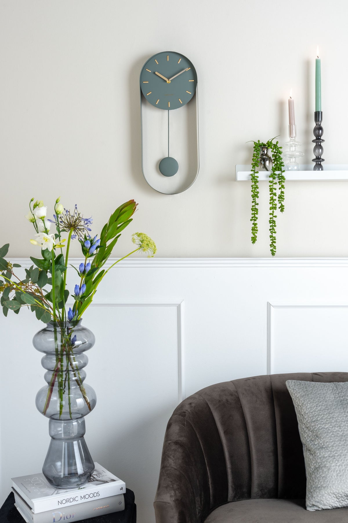Karlsson wandklok Pendulum Charm, staal jungle groen, sfeerfoto: aan lichte wand met lambrisering, bloemen