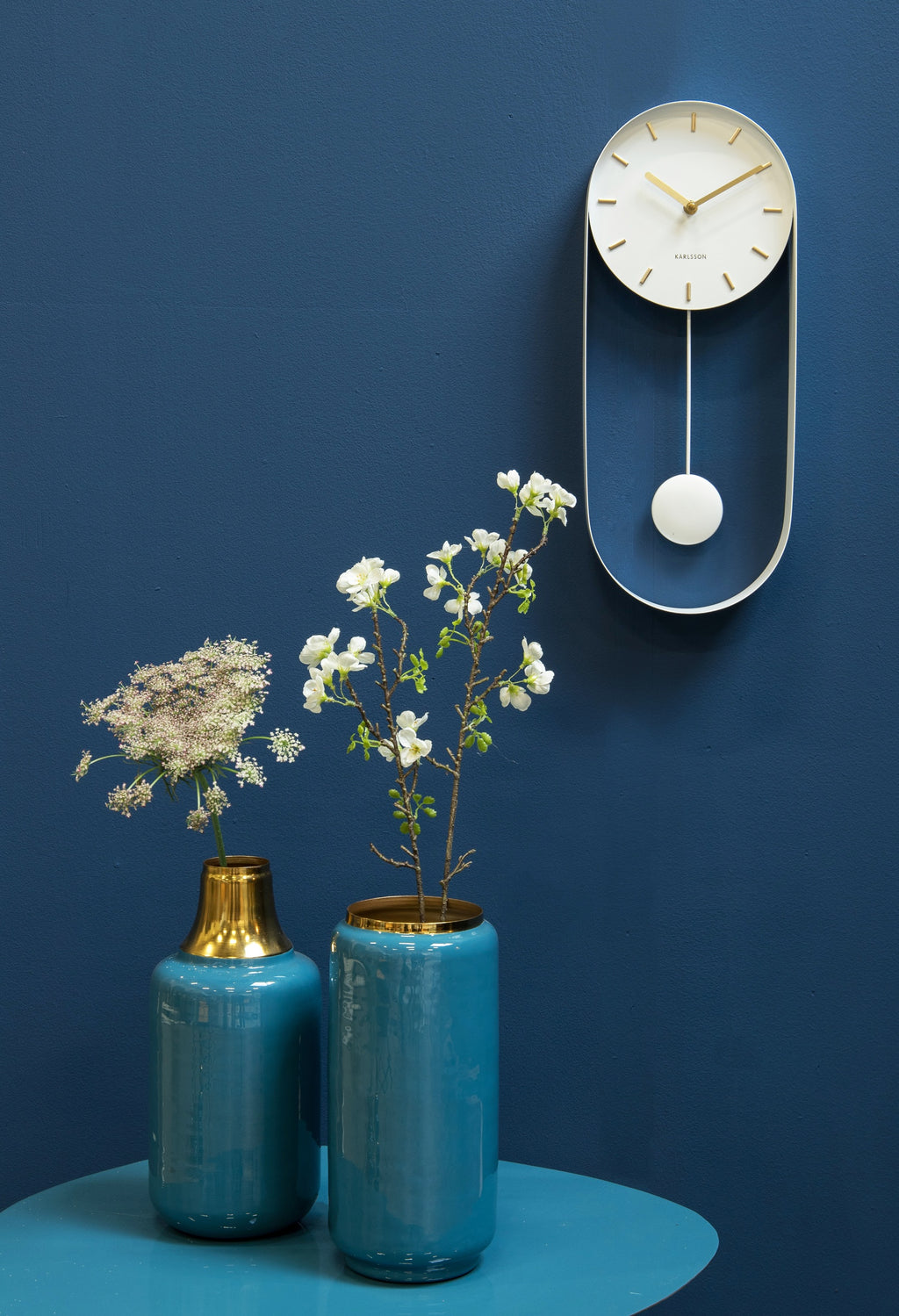 Karlsson wandklok Pendulum Charm, staal wit, sfeerfoto: aan blauwe wand naast blauwe vazen met bloesem