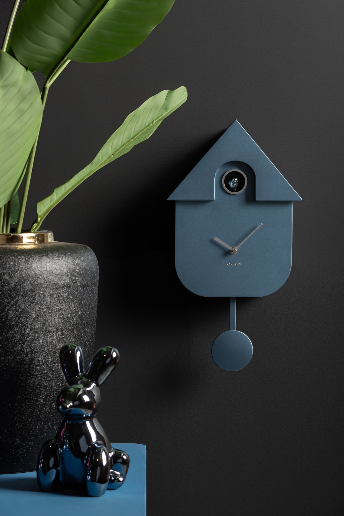 Karlsson koekoeksklok Modern Cuckoo, donkerblauw abs metallic, sfeerfoto: aan donkere muur naast plant en glanzend konijn