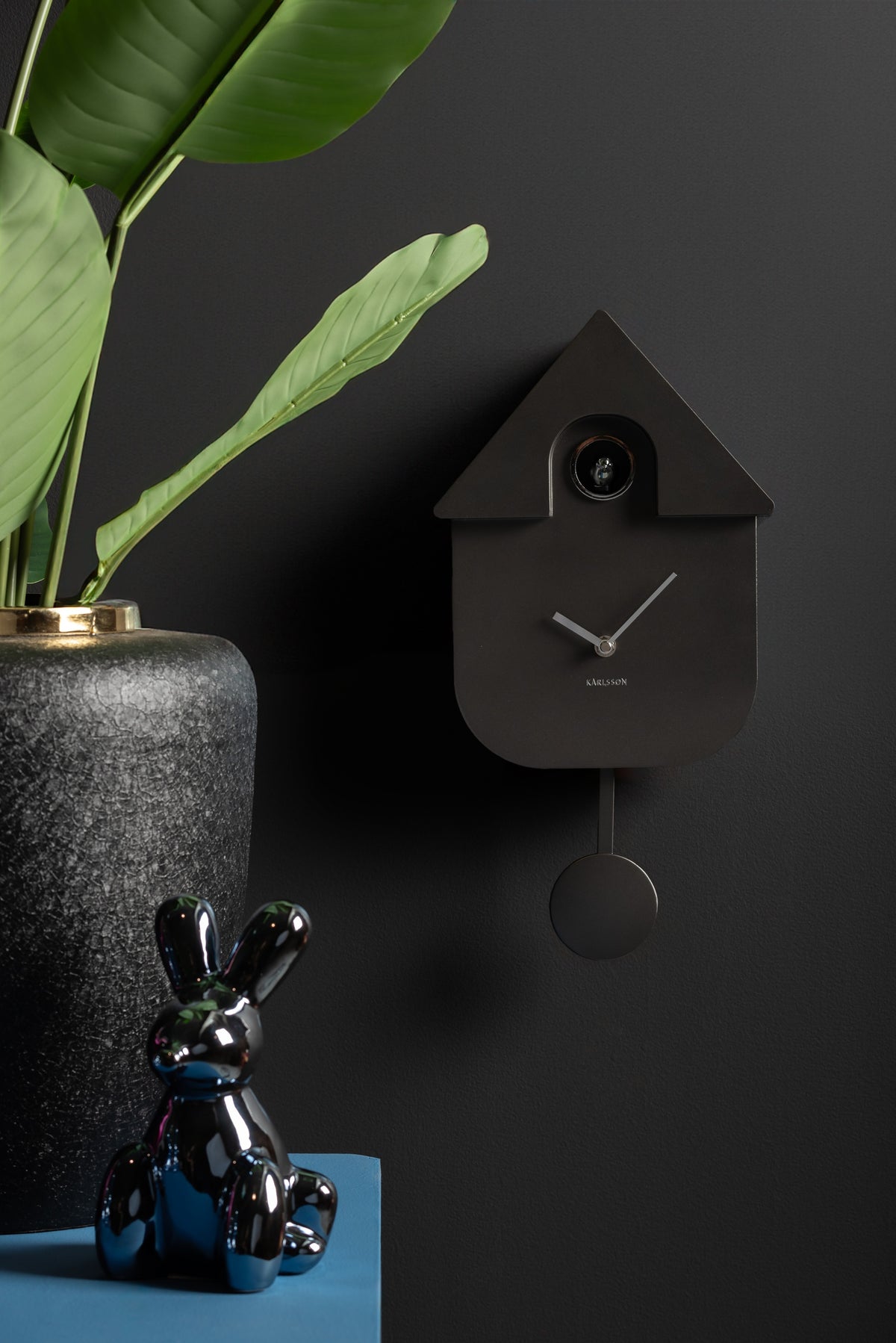 Karlsson koekoeksklok Modern Cuckoo, donkergrijs abs metallic, sfeerfoto: aan donkere muur naast plant en glanzend konijn
