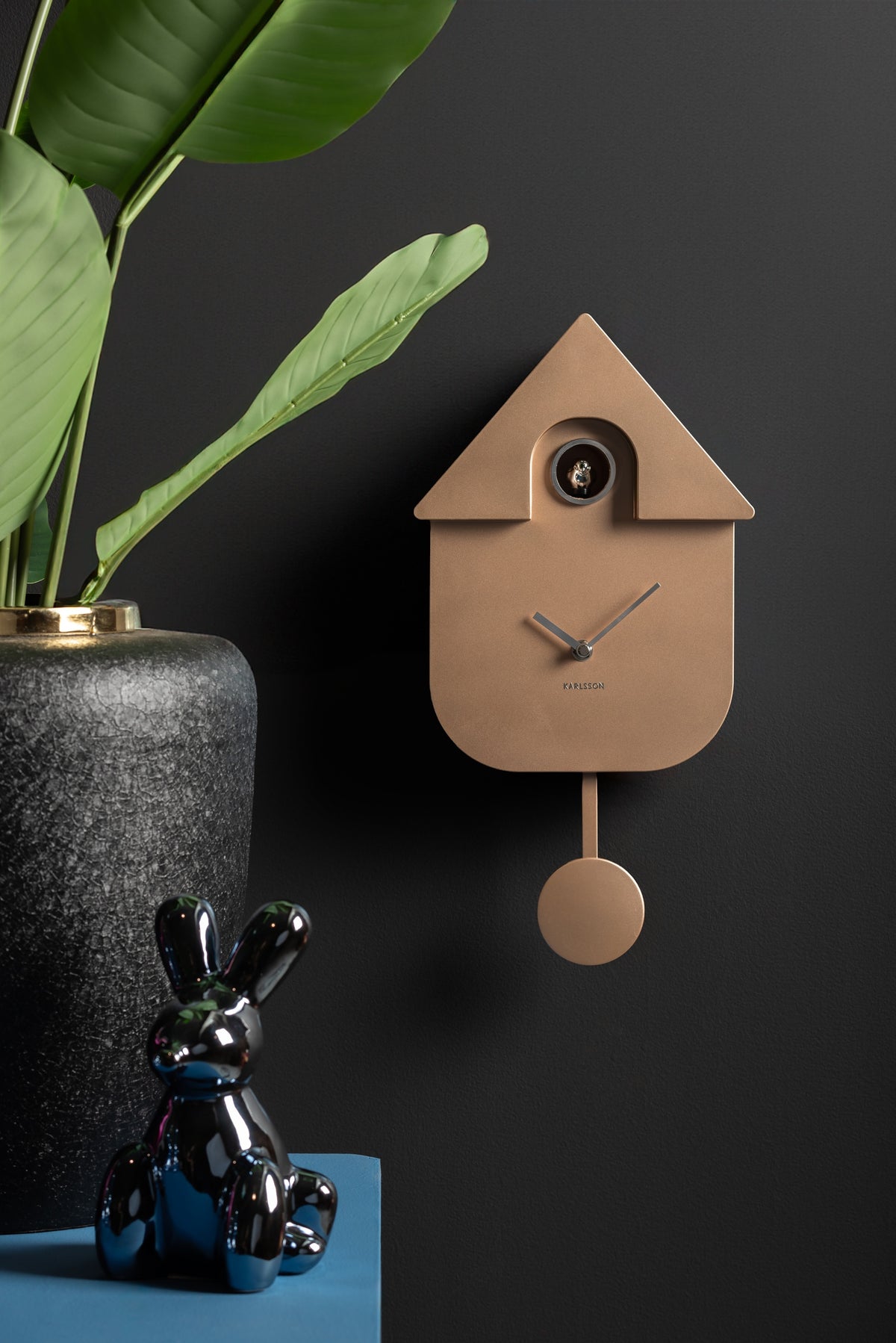 Karlsson koekoeksklok Modern Cuckoo, rosé goud abs metallic, sfeerfoto: aan donkere muur naast plant en glanzend konijn