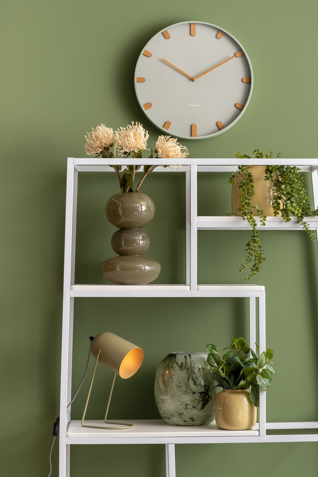 Karlsson wandklok Grace, groen, sfeerfoto: aan groene wand boven wit stalen wandrek met vazen, planten en lampje
