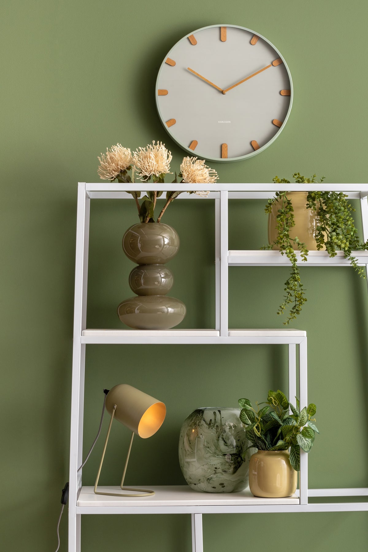 Karlsson wandklok Grace, groen, sfeerfoto: aan groene wand boven wit stalen wandrek met vazen, planten en lampje