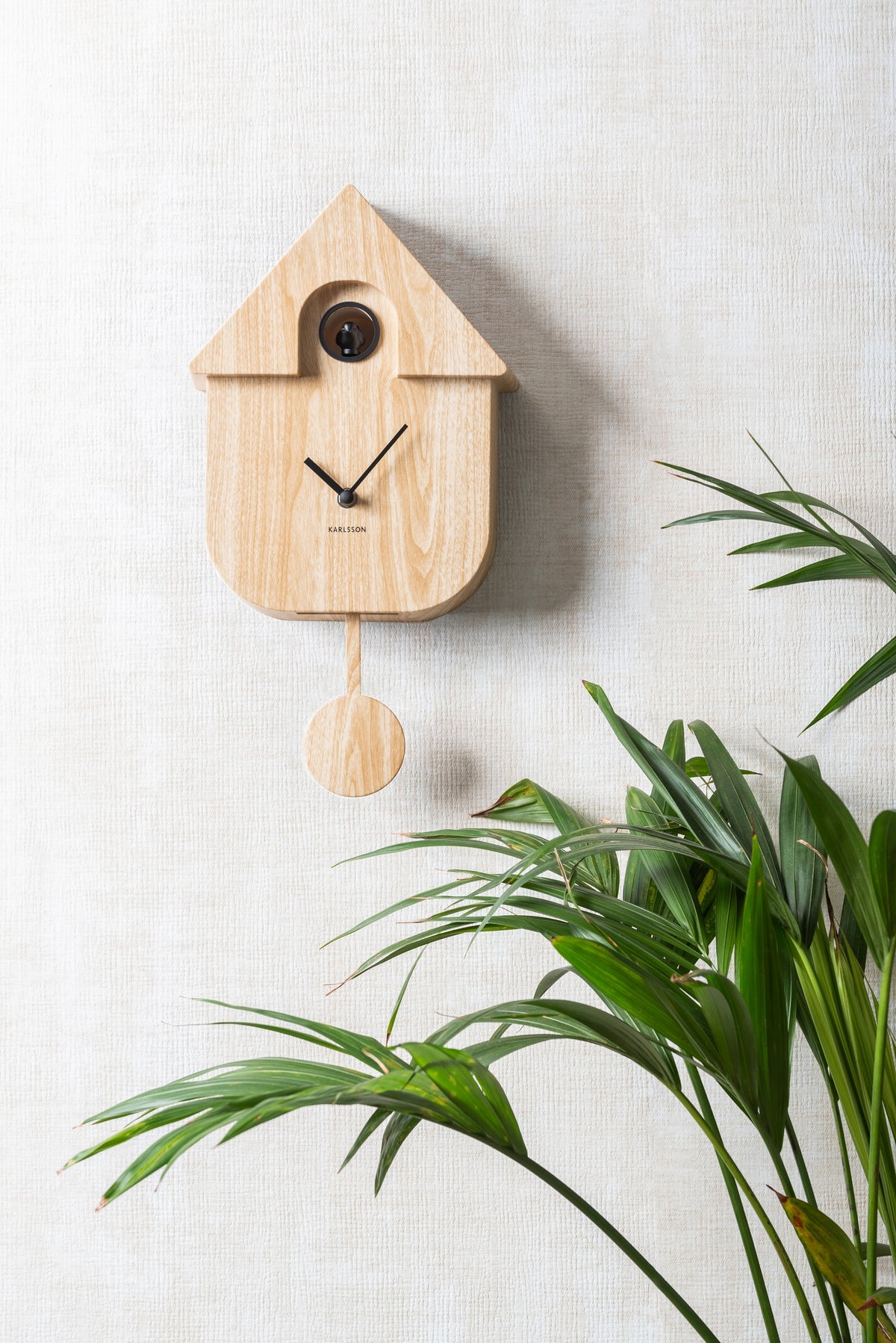 Karlsson koekoeksklok Modern Cuckoo, licht hout print, sfeerfoto: aan lichte wand naast palmbladeren
