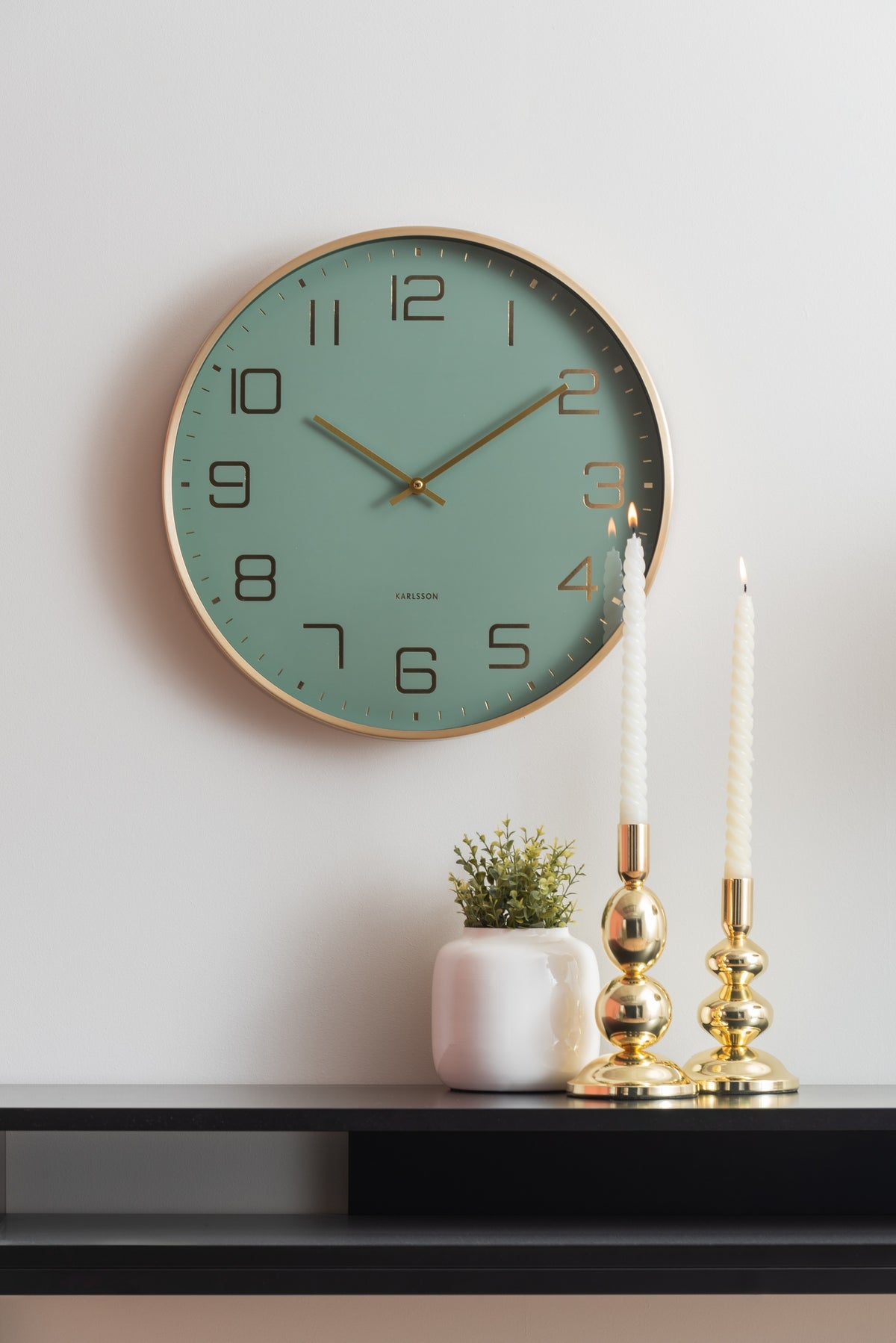 Karlsson wandklok Elegance Glow, groen, sfeerfoto: aan witte muur boven zwart dressoir met gouden kandelaars en plantje