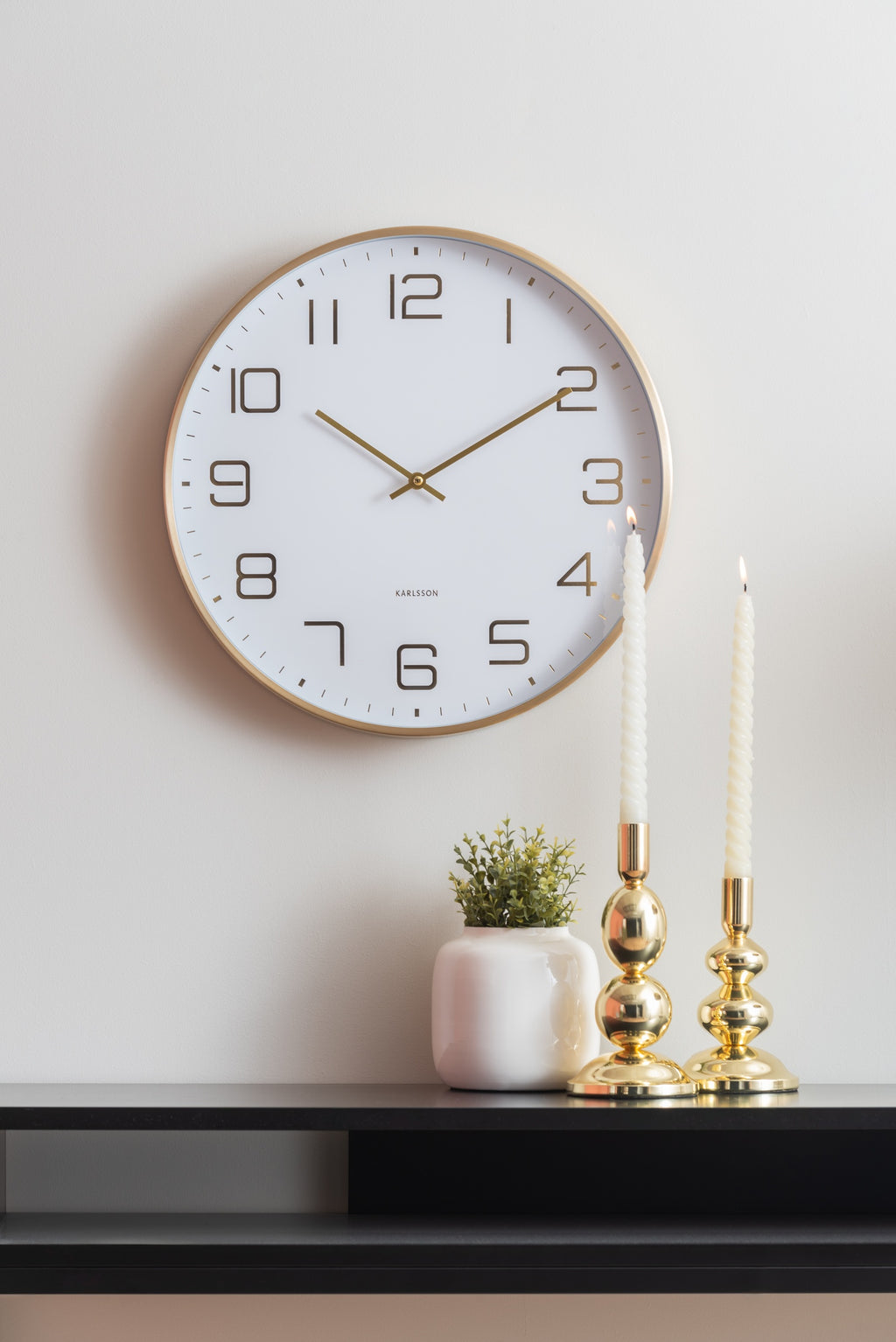 Karlsson wandklok Elegance Glow, wit, sfeerfoto: aan witte muur boven zwart dressoir met gouden kandelaars en plantje