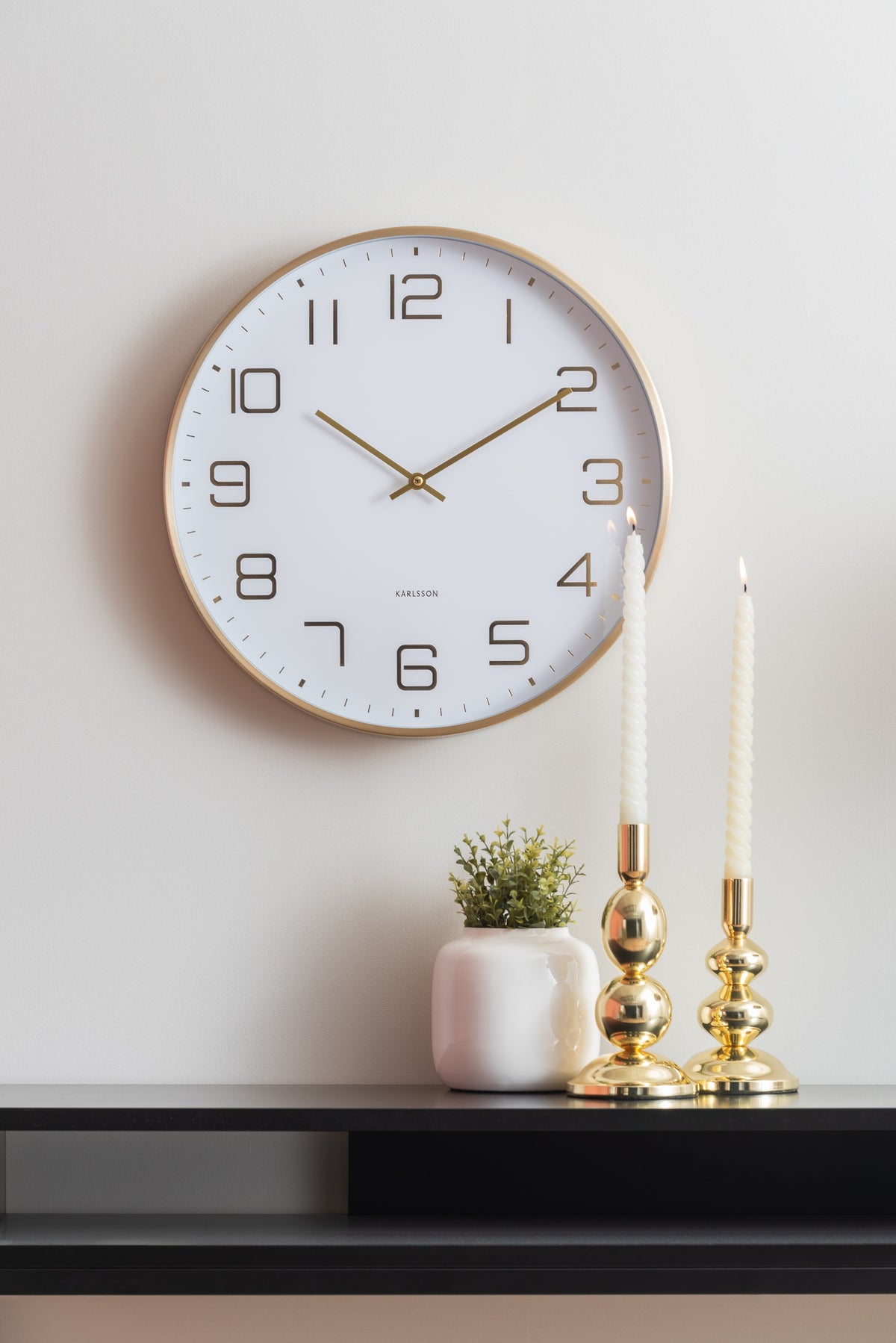 Karlsson wandklok Elegance Glow, wit, sfeerfoto: aan witte muur boven zwart dressoir met gouden kandelaars en plantje