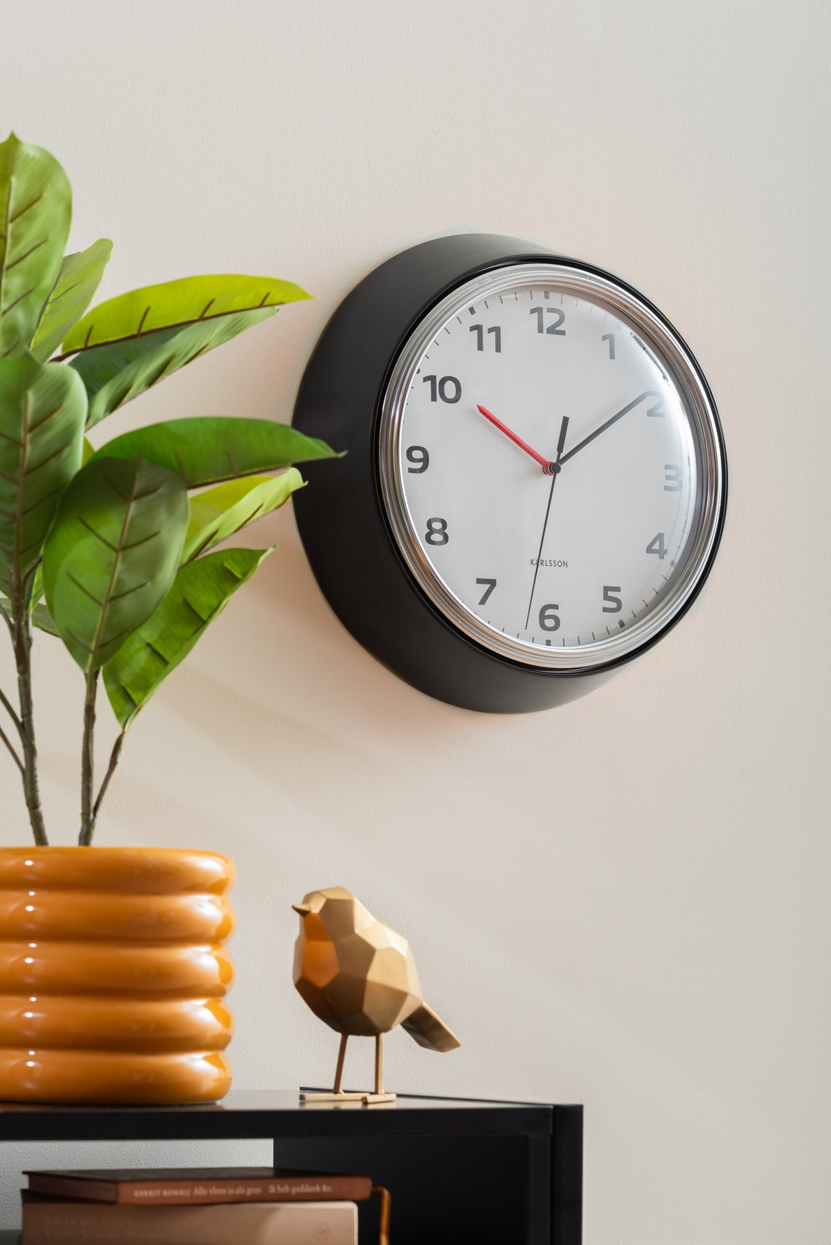 Karlsson wandklok Modern Retro, zwart, sfeerfoto: aan lichte muur naast plant en gouden vogeltje op kast