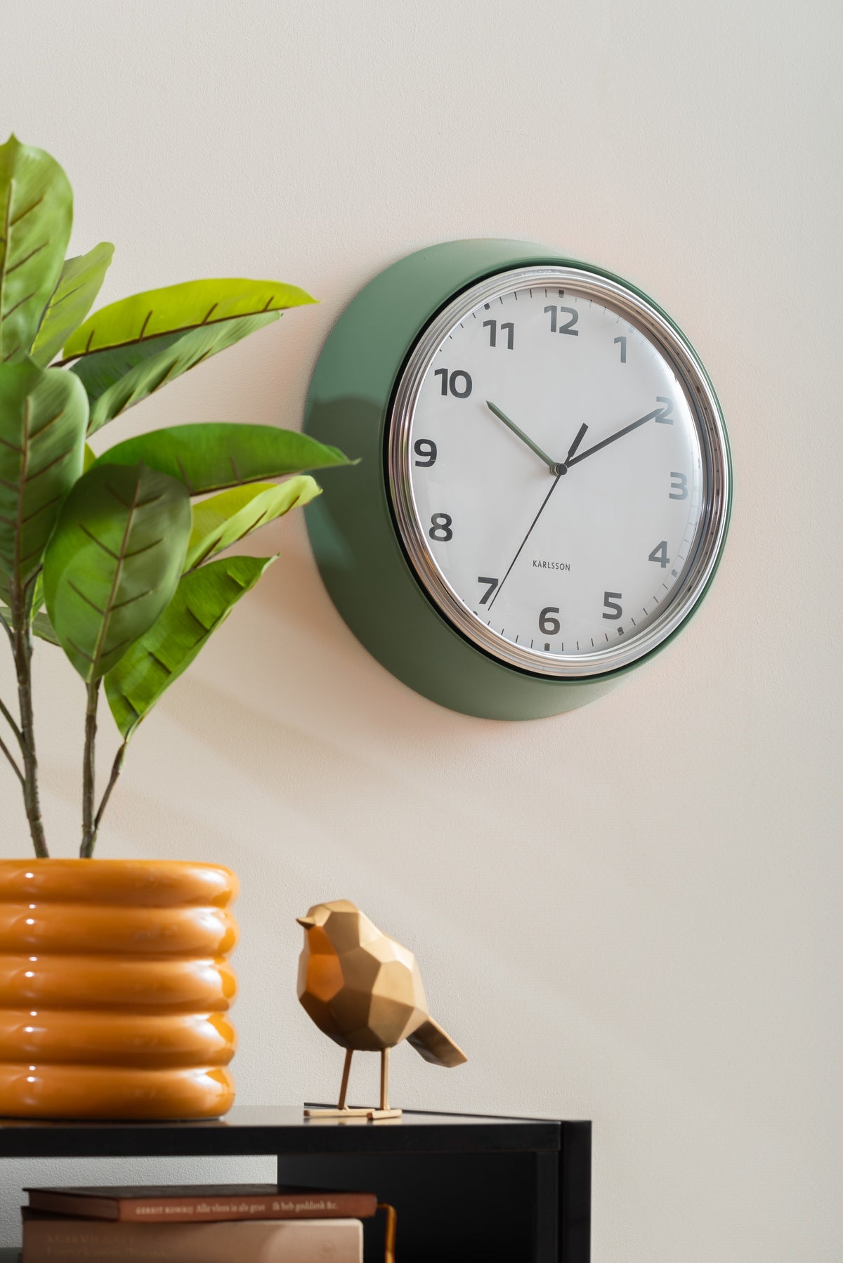 Karlsson wandklok Modern Retro, groen, sfeerfoto: aan lichte muur naast plant en gouden vogeltje op kast