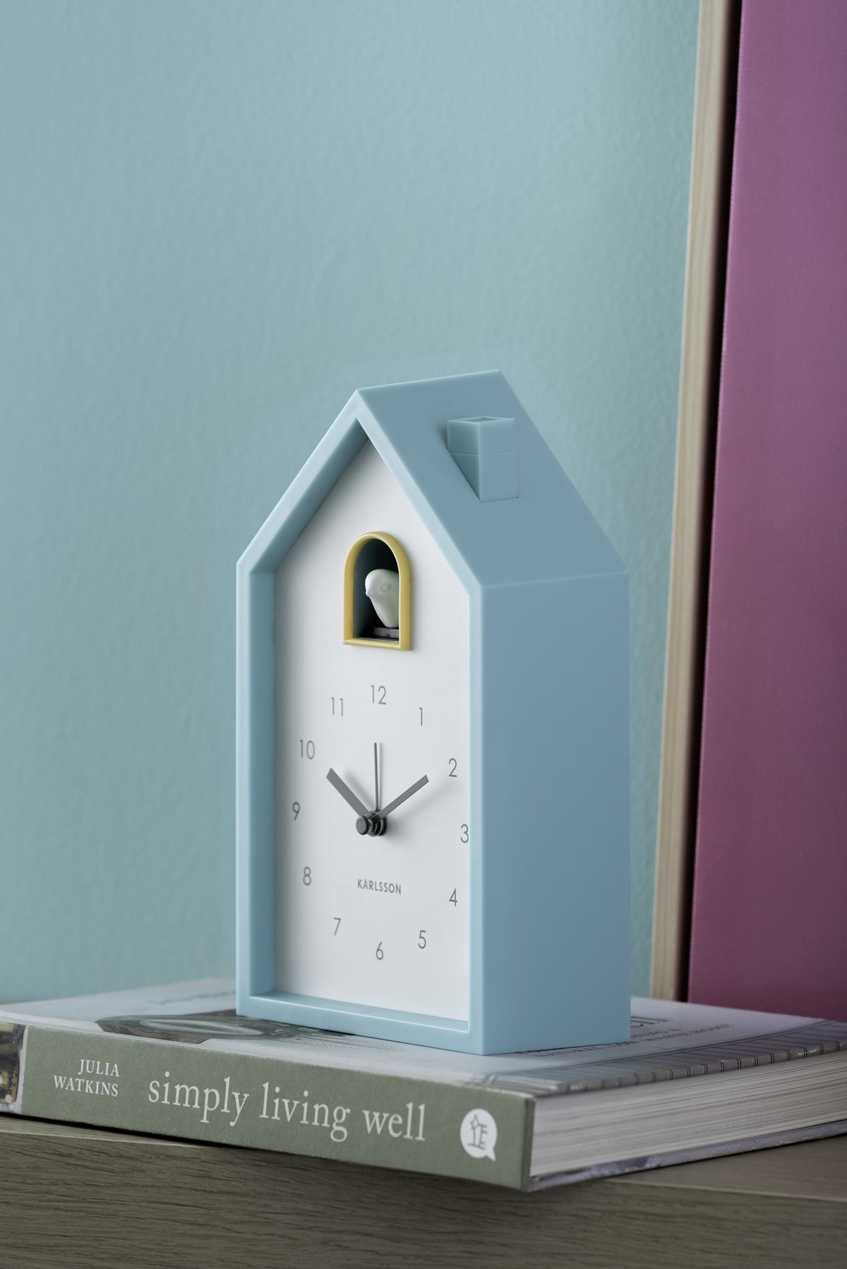 Karlsson wekker Modern Cuckoo, turquoise blauw, sfeerfoto: op boek tegen turquoise muur met roze paneel