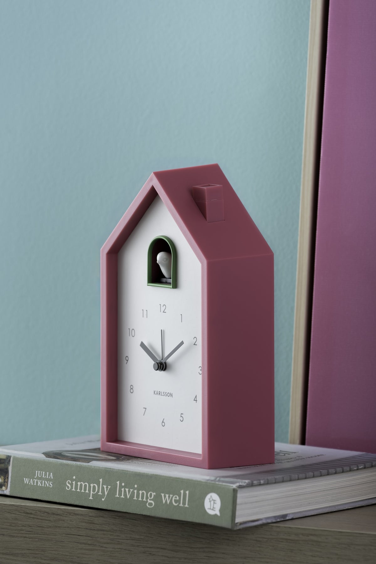 Karlsson wekker Modern Cuckoo, helderroze, sfeerfoto: op boek tegen turquoise muur met roze paneel
